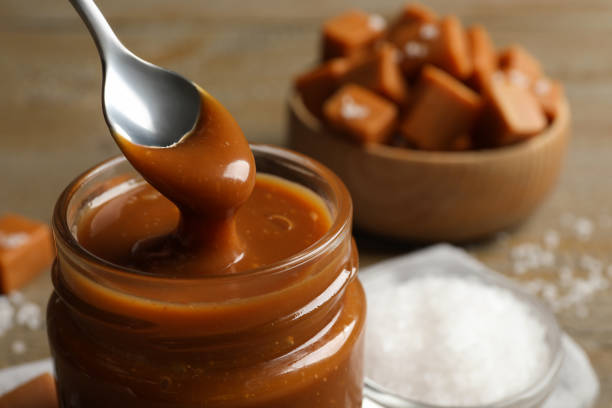Taking yummy salted caramel with spoon from glass jar at table, closeup. Space for text