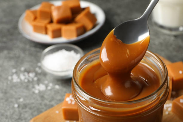 istock Taking yummy salted caramel with spoon from glass jar at table, closeup. Space for text 1421199044