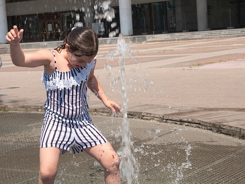 A happy little girl is bathing in a fountain spouting high jets of water from the ground, dancing to music. Summer entertainment.