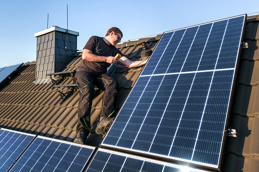 A man sits on the roof of a house while checking the photovoltaic system with a multimeter.