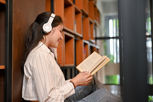 Relaxed and happy young Asian female in a bookstore listening to music through headphones while focused on reading a book. Education and lifestyle concept