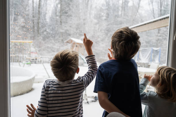 Three siblings, brothers and sister, looking out the window amazed by the snowy winter nature stock photo