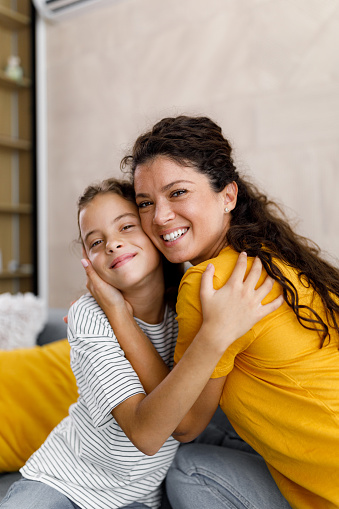 Young happy mother and her little girl embracing in the living room and looking at camera.