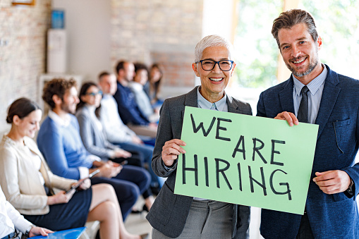 Happy business managers holding announcement message with  the sign 'We are hiring' and looking at camera. Candidates are in the background.