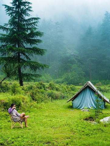 July 7th 2021. A man sipping on a hot tea cup in the meadows with a camp amid deodar tree forest and mountains in the background. Uttarakhand India.