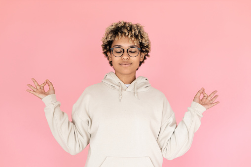 African American woman meditating in studio. Female model in hoodie and glasses with closed eyes. Portrait, studio shot, gesture concept