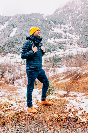 Winter hike on snow mountain young happy hiker man walking. Europe travel adventure trek in nature landscape. Young bearded person wearing yellow hat, blue jacket, jeans for cold weather and bag.