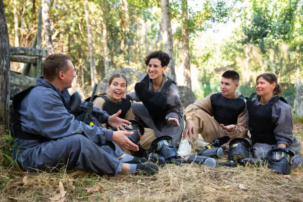 Positive paintball players sitting on ground during break between rounds, smiling and chatting between each other.