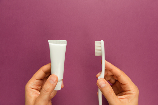 Hands are holding white toothpaste and white toothbrush on pink background.