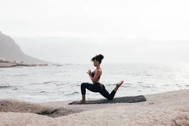 Photo of Side view of fit woman practicing yoga on mat by ocean. Female meditating at evening with folded hands.