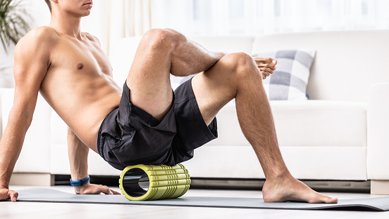 A young man massages the gluteal muscle with a foam roller.