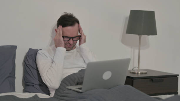 istock Casual Man having Headache while Working on Laptop in Bed 1421247439