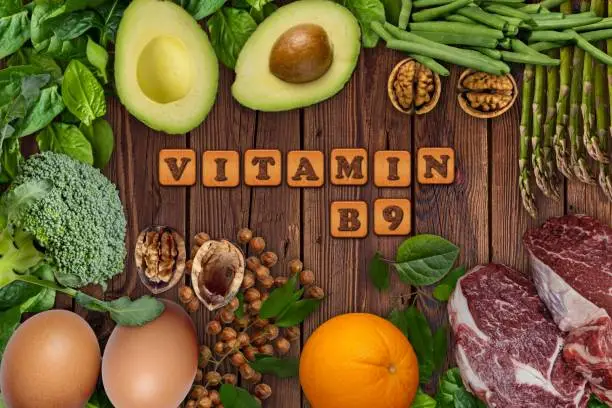 Photo of Natural sources of vitamin B9 (folic acid) . Foods as Avocado, eggs, orange, broccoli, liver, asparagus, beans, spinach, nuts and seeds  on wooden table.