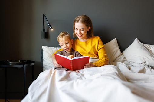 Cheerful family: woman and boy son smiling and reading fairy tale while relaxing on bed  at home