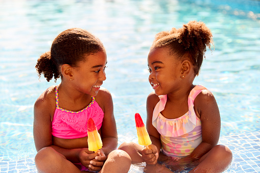 Family On Summer Holiday With Two Girls Eating Ice Lollies By Swimming Pool