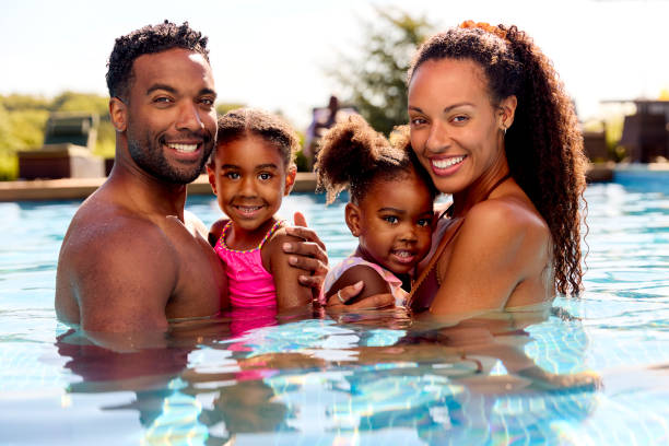 Portrait Family On Summer Holiday With Two Girls Being Held In Swimming Pool By Parents stock photo