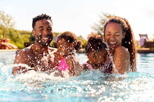 Family On Summer Holiday With Two Girls Being Held In Swimming Pool By Parents And Splashing stock photo