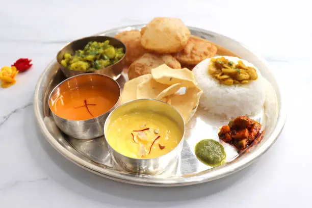 Indian vegetarian lunch or dinner Thali includes Aloo ki sabji, dal rice, Puri bhaji, Shrikhand or Srikhand, Aamras, papad, pickle, and chutney. Indian food is served in a Silverplate or thali. copy.