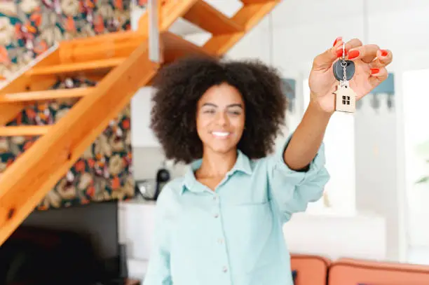 Selective focus at keys with cute keychain little house showing by young happy woman, excited female student moved in own dwelling, african-american relocated, moving into a new house, got a mortgage