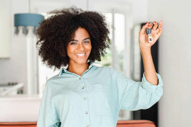 Happy overjoyed young woman with afro hairstyle holding keys with keychain in form of little house indoors. Smiling multiracial woman holding keys from new property, happy buyer of own estate stock photo