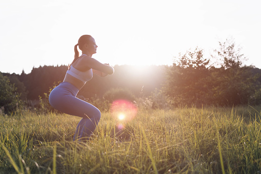 a woman of light appearance in blue cleats and a white tank top is engaged in fitness in the park standing on the grass on a mat, the concept of a healthy lifestyle. silhouette of a sports girl at sunset, endurance. High quality photo