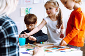 Two beautiful little girls standing at table with pencils in bright classroom. Little boy sitting, playing board games.