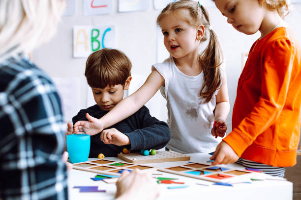 dwie piękne dziewczynki stojące przy stole z ołówkami w jasnej klasie. mały chłopiec siedzi, grając w gry planszowe. - teacher child preschool mathematics zdjęcia i obrazy z banku zdjęć