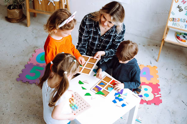 vue de dessus d’un groupe d’enfants d’âge préscolaire. jeune femme blonde aidant les élèves à jouer à des jeux de société, collectionnant des figures géométriques. - early childhood education photos et images de collection