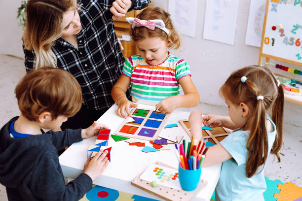 Portrait of young pretty woman sitting at table with pupils children playing. Teacher touching headband of little girl. Portrait of young woman sitting at table with colored pencils, board games with pupils children playing. Teacher touching pink and white headband of little girl collecting puzzles in bright classroom. montessori stock pictures, royalty-free photos & images