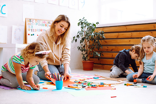 Portrait of young smiling woman teacher sitting near colorful pencils, different toys with wonderful children, checking looking at painting on red paper in bright classroom. Education, forwardness.