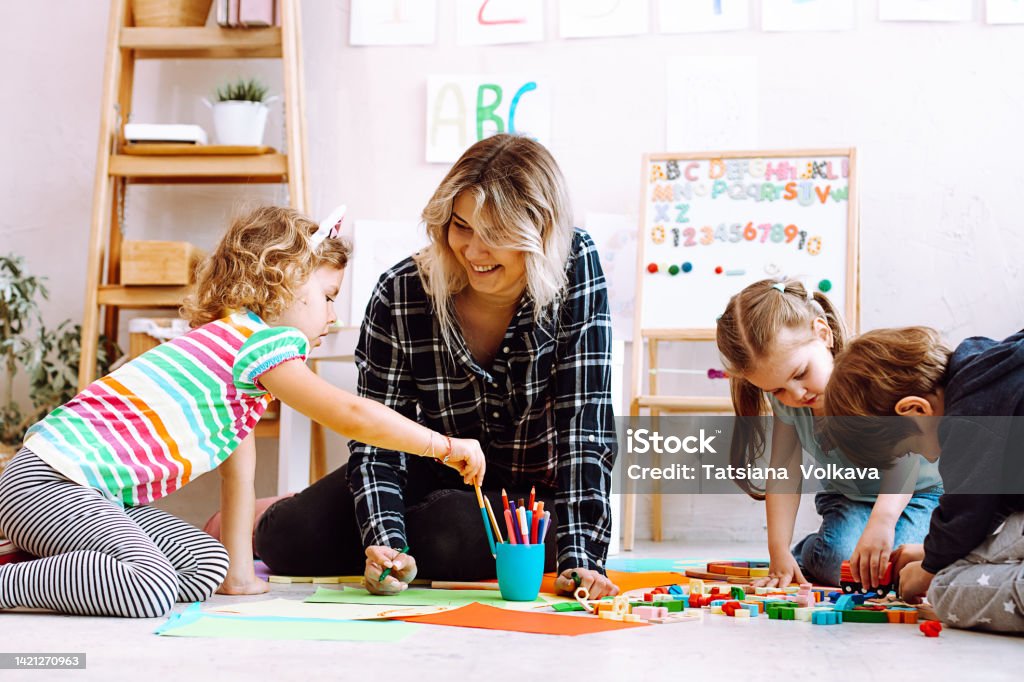 Portrait of young joyful woman teacher sit with children, looking at serious little girl taking pencil in classroom. Portrait of young joyful woman teacher sitting near colored pencils, different toys with playing children, looking at serious little girl taking pencil in bright classroom. Education, forwardness. Preschool Stock Photo