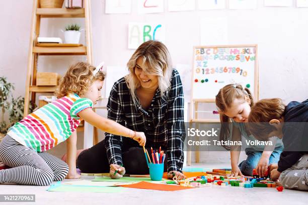 Portrait Of Young Joyful Woman Teacher Sit With Children Looking At Serious Little Girl Taking Pencil In Classroom Stock Photo - Download Image Now