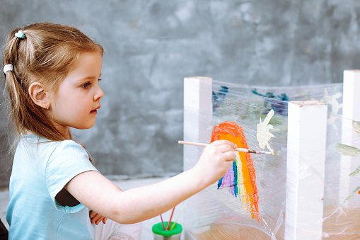 Side view of wonderful little girl sitting on art lesson, painting drawing white sun near rainbow with brush on stretch film wrapping around turned table legs in grey classroom. Early development.