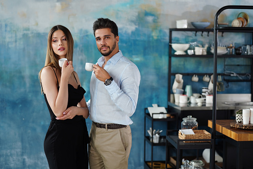 Attractive elegant couple enjoying their early morning coffee, standing with espresso in hands in designer kitchen. Real people luxury lifestyle.