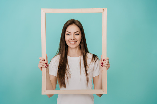 Young Italian woman with long hair in a white T-shirt stands on a turquoise background, holds a wooden frame for a picture, smiles, looks at the camera. Mockup, Creative people, idea, youth.