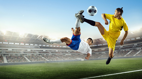 Football match. Athletic men, soccer football players fight for the ball at crowded 3d stadium during sport match over blue cloudy sky background. Sport competitions.