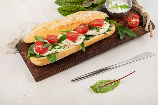 Preparing a fresh crusty baguette with cherry tomatoes, arugula and mozzarella cheese on a wooden board with sauce. White background. Top view.