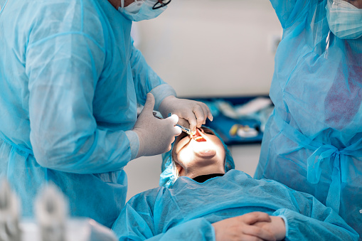 Male dentist wearing face mask and protective clothes doing treatment to unrecognized patient.