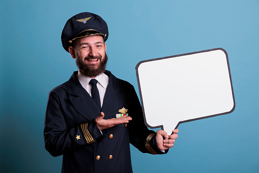 Smiling airplane aviator holding white blank speech bubble, communication frame, discussion mock up. Plane pilot in aviation uniform standing near empty message balloon with place for text