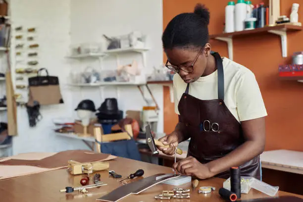 Portrait of black of female artisan punching holes in handmade leather belt at workshop, copy space