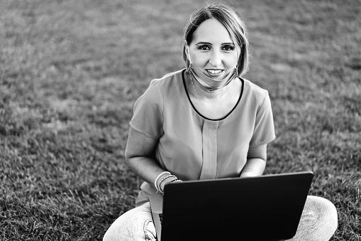 Woman 40 years old working outdoors with laptop during coronavirus outbreak - Smiling female entrepreneur sitting on lawn with personal computer while looking camera - Remote work concept into nature