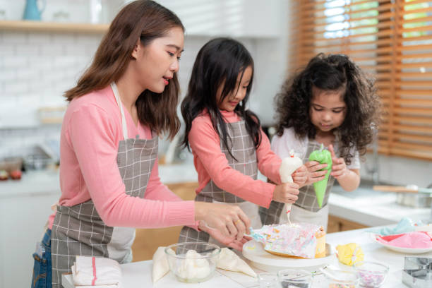 junges kleines mädchen, das in der thekenbäckerei in der modernen küche zu hause steht. glückliche lächelnde eltern genießen das wochenende. kind und mutter in schürze dekorieren creme auf hausgemachtem kuchen. lernkonzept - school classroom child back to school stock-fotos und bilder