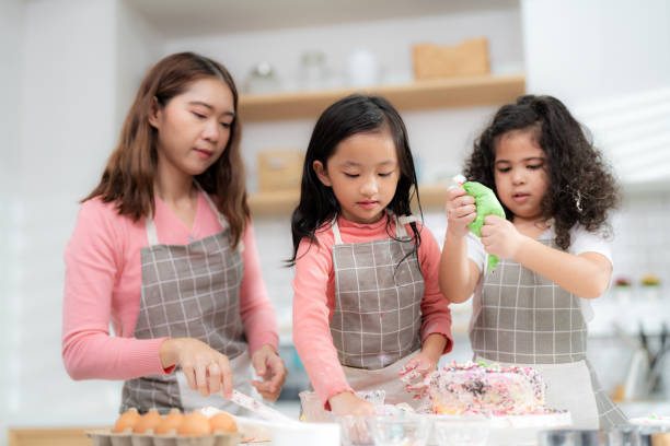 junges kleines mädchen, das in der thekenbäckerei in der modernen küche zu hause steht. glückliche lächelnde eltern genießen das wochenende. kind und mutter in schürze dekorieren creme auf hausgemachtem kuchen. lernkonzept - school classroom child back to school stock-fotos und bilder