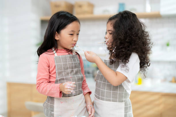 junges kleines mädchen, das in der thekenbäckerei in der modernen küche zu hause steht. viel spaß beim lächeln und genießen am wochenende. zwei kinder in schürze füttern sich gegenseitig mit kuchen. hausgemachtes bäckereikonzept - school classroom child back to school stock-fotos und bilder