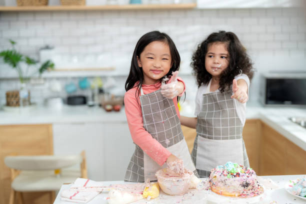 junges kleines mädchen, das in der thekenbäckerei in der modernen küche zu hause steht. viel spaß beim lächeln und genießen am wochenende. zwei kinder tragen schürze und daumen hoch. hausgemachte bäckerei und lernklassenkonzept - school classroom child back to school stock-fotos und bilder