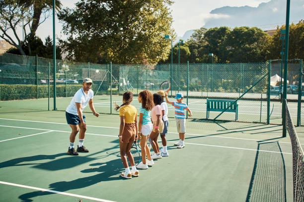 Tennis sports teacher coaching children, students or kids on a tennis court for sport education. Fitness, exercise and workout as coach teaching a training game to a young learning student class Tennis sports teacher coaching children, students or kids on a tennis court for sport education. Fitness, exercise and workout as coach teaching a training game to a young learning student class kids tennis camp stock pictures, royalty-free photos & images