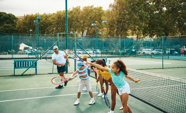 Tennis coach teaching group of children on a court during exercise, training or workout to improve skills and swing. Kids with racket and ball practicing for a game or match during a sport coaching Tennis coach teaching group of children on a court during exercise, training or workout to improve skills and swing. Kids with racket and ball practicing for a game or match during a sport coaching sports training camp stock pictures, royalty-free photos & images