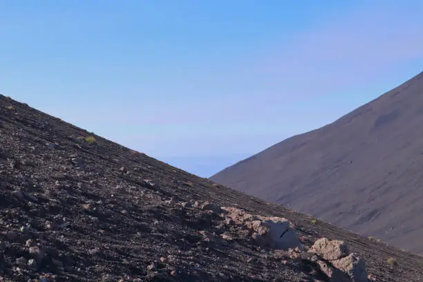 Photo of Rocky mountainsides of Mnt Etna forming two dark triangles in large contrast with early morning sun and lightly clouded blue sky