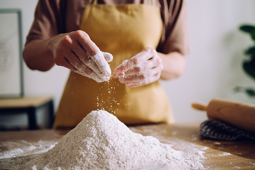 Close-up of unrecognizable woman kneading bread dough on wooden kitchen counter. She is preparing sourdough bread sprinkling flour over the dough.