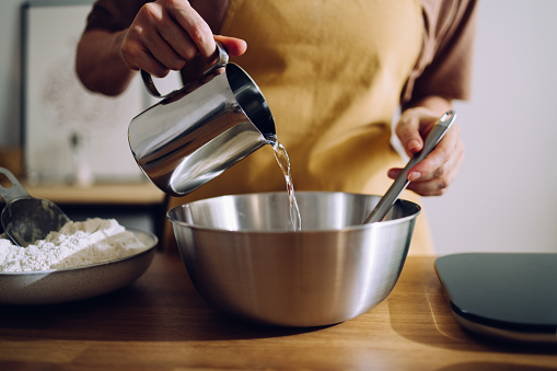Unrecognizable woman holding a metal ewer and pouring water into the dough mixture and mixing it with the spatula. She is getting ready to bake.